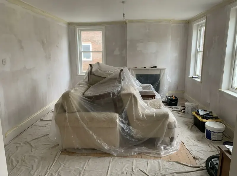 Woonkamer waarvan de meubels naar het midden geschoven zijn en met folie afgedekt