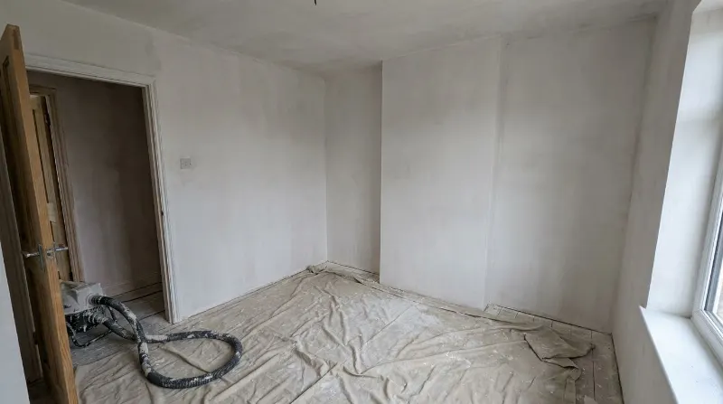 Slaapkamer met pas bepleisterde witte muren, folie op de vloer en de slang van de pleistermachine bij de deur